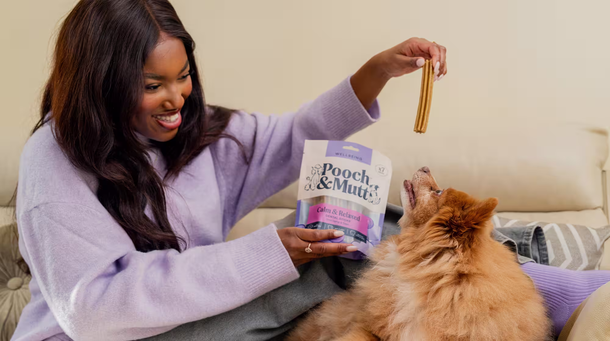 Sat on a sofa, a dog owner dangles a dental chew above dog who is looking up trying to reach it