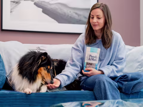 A dog owner feeds their dog from a packet of Pooch & Mutt Meaty Treats whilst sat on the sofa.